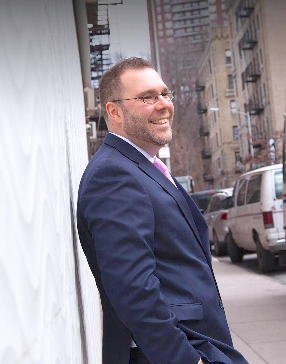 Matthew Bizzarro smiling, leaning on a white wall on a New York City street